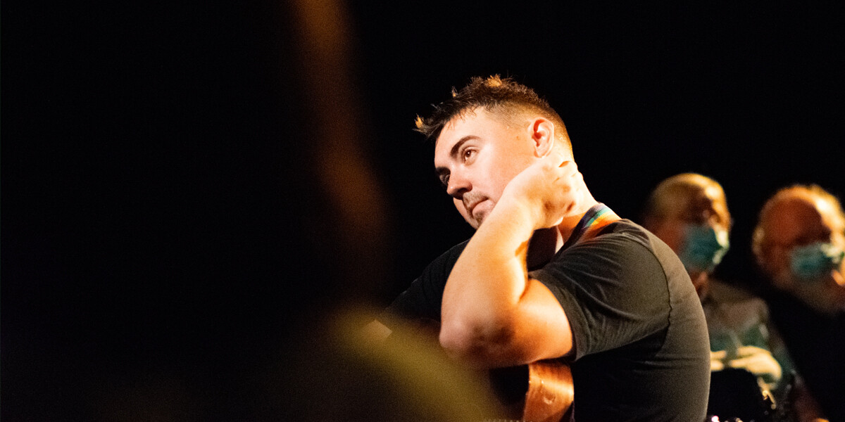 A man holding a guitar sat watching another performer (off camera)