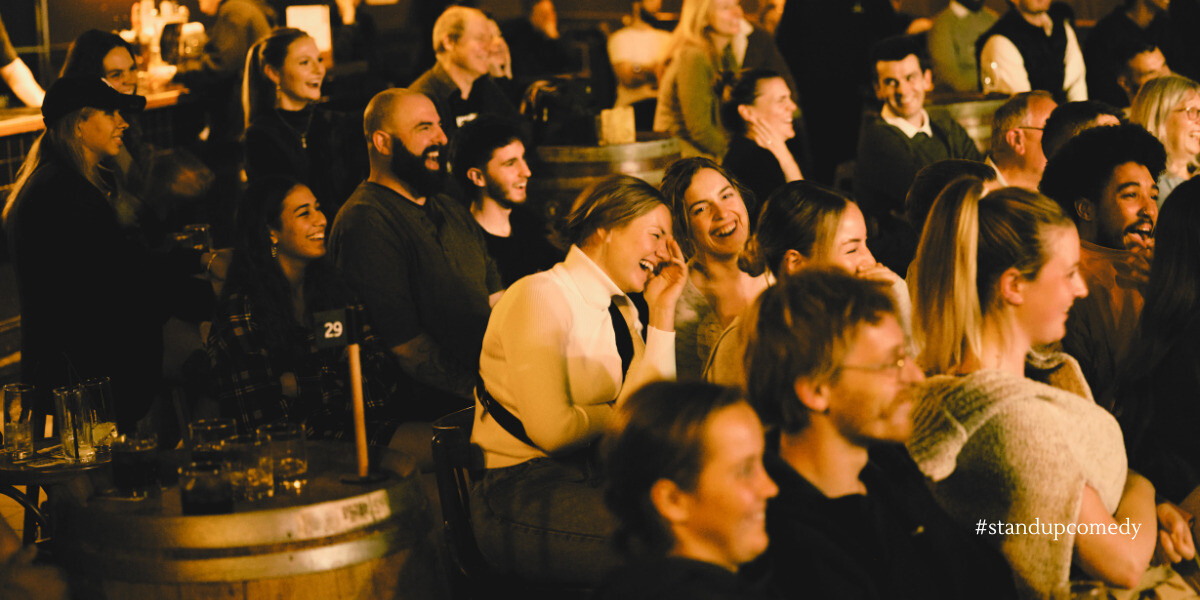 BOOMERS AND ZOOMERS: Comedy Showcase!–Audience laughing and enjoying a live stand-up comedy show in a warmly lit venue. People sit around wooden tables and barrels with drinks, smiling and reacting to the performer off-camera. The scene captures the lively, communal atmosphere of a packed comedy night. The hashtag #standupcomedy appears in the corner.