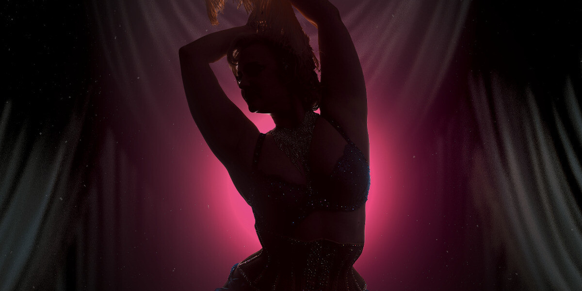 Teaser–Woman lit by silhouette with her arms in the air wearing a corset, bra and feather in her hair. Background is pink.