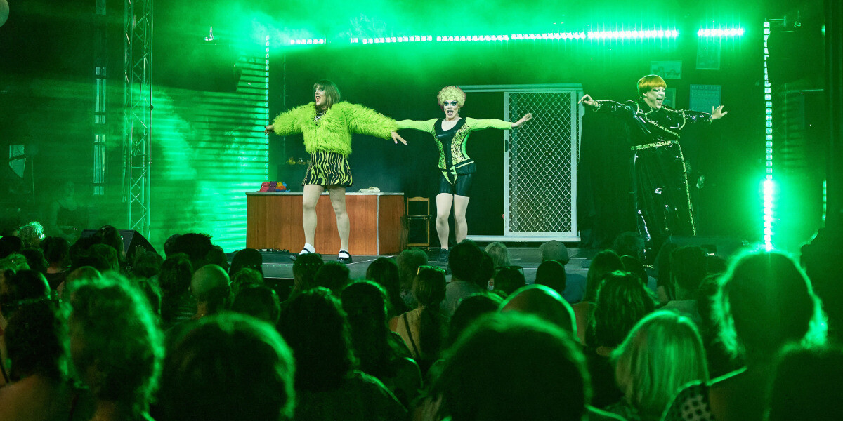 Four performers dance in formation on a colourful stage under rainbow lighting, facing a packed audience. From left to right: green mini dress, purple top with white pants, brown jacket and trousers, and a pink two-piece with “NWD” printed on the crop top.
